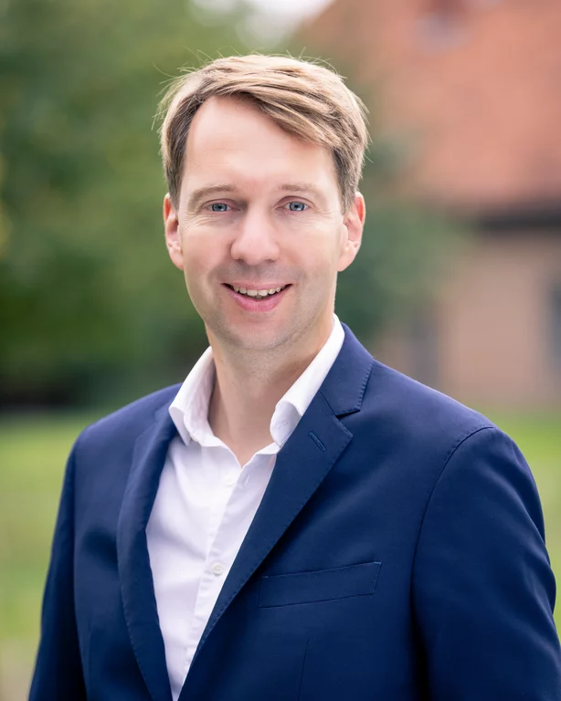 Das Porträt zeigt Jan Dingeldey in einer klassischen, vertrauenserweckenden Kameraperspektive als Brustbild. Er blickt mit einem freundlichen, offenen Lächeln direkt in die Kamera, was eine unmittelbare Verbindung zum Betrachter herstellt. Sein Erscheinungsbild ist professionell und modern: Er trägt ein dunkelblaues Sakko mit dezenter Textur über einem reinweißen Hemd, das am Kragen modisch offen steht – ein Look, der Kompetenz mit Nahbarkeit vereint. Der Hintergrund der Aufnahme ist bewusst in einer sanften Unschärfe (Bokeh-Effekt) gehalten. Man erkennt vage natürliche Grüntöne von Bäumen oder Sträuchern sowie die warmen Nuancen eines Backsteingebäudes oder eines Ziegeldachs. Diese Wahl des Hintergrunds lässt das Porträt lebendig und authentisch wirken, ohne vom Hauptmotiv abzulenken.